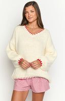 Girl is styled in an oversized cream chunky knit sweater featuring red contrast stitching along the V-neck and cuffs. She wears it with red and white striped shorts and beige shearling-lined slippers over white socks with red heart details.