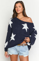 Model is wearing a navy blue chunky knit sweater adorned with large white knitted star patterns on the body and sleeves. The sweater has a wide neckline and is paired with navy and white horizontally striped pants.